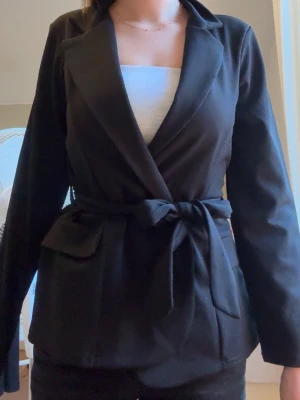 Svart kavaj med knytskärp - Stilren svart kavaj med klassisk krage och knytskärp i midjan. Kavajen har långa ärmar, två fickor framtill och rak passform. Perfekt för en trendig och snygg look. Aldrig använd