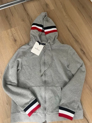 Grå Moncler hoodie med dragkedja - Passform M. Snygg grå hoodie från Moncler med dragkedja framtill och huva med dragsko. Huva och ärmslut har röd, vit och marinblå randdetalj som ger en sportig vibe. Perfekt för kallare sommardagar.