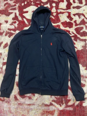 Marinblå hoodie från Polo Ralph Lauren - Snygg marinblå hoodie från Polo Ralph Lauren med dragkedja och huva. Klassisk röd logga broderad på bröstet och två fickor framtill. Modellen är 'Custom Fit' och materialet är mjuk bomull, perfekt för en avslappnad stil.