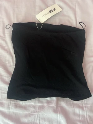 Svart bandeau tubtopp från FBSister - Säljer en svart bandeau tubtopp från new yorker. Toppen är axelbandslös och har en tight passform som sitter snyggt på kroppen. Perfekt att styla med jeans eller kjol för en clean look.