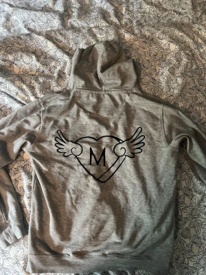Mira Paris grå hoodie  - Säljer en grå hoodie med huva och magficka. På ryggen finns ett stort tryck med ett hjärta, bokstaven M och vingar. Hoodien har dragsko i huvan och är gjord i mjukt sweatshirtmaterial, perfekt för en chill och avslappnad stil. NY PRIS RUNT 1700kr 💰