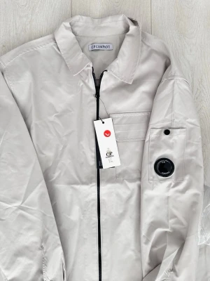 Beige overshirt från C.P. Company - Stilren beige overshirt från C.P. Company med dragkedja framtill, klassisk krage och bröstficka. Ärmen har en ikonisk linsdetalj och ficka med knapp. Tillverkad i ett slätt, lätt material som ger en modern look. Perfekt lager på lager-plagg för dig som gillar streetwear.