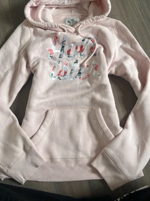 Ljusrosa hoodie från Hollister - Säljer en ljusrosa hoodie från Hollister med stor magficka och huva med dragsko. Framsidan har ett färgglatt Hollister-tryck i blommigt mönster. Mjuk insida i fleece och ribbade muddar vid ärmar och nederkant. Perfekt för en chill och avslappnad stil.