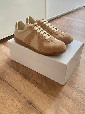 Maison margiella - Snygga beige sneakers med ovandel i mocka och släta läderpaneler. Klassisk låg modell med vita snören och gummisula. Perfekta för dig som gillar stilrena och neutrala färger. Passar till det mesta och ger en clean look.