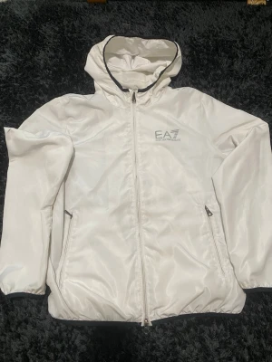 Vit vindjacka från EA7 Emporio Armani - Snygg vit vindjacka från EA7 Emporio Armani med huva och dragkedja framtill. Jackan har två sidofickor med dragkedja och meshfoder på insidan för extra komfort. EA7-logga på bröstet och elastiska muddar vid ärmslut och nederkant.
