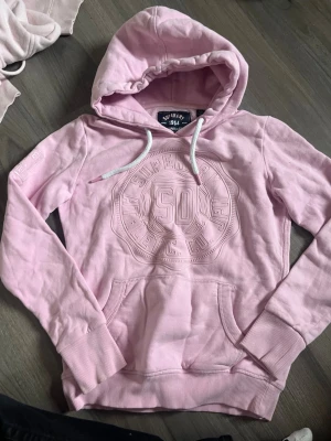 Ljusrosa hoodie från Superdry - Säljer en ljusrosa hoodie från Superdry med broderad logga och text på bröstet. Hoodien har huva med vita dragsnören, magficka och ribbade muddar vid ärmar och nederkant. Tillverkad i mjuk bomullsblandning för extra komfort.