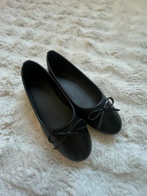 Svarta ballerinaskor med rosett - Stilrena svarta ballerinaskor med rund tå och dekorativ rosett framtill. Skorna har en klassisk och enkel design i ett glansigt material som ger en snygg look. Perfekta att matcha till olika outfits för en trendig touch.
