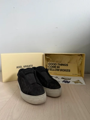 Svarta slip-on sneakers Axel Arigato - Säljer ett par svarta slip-on sneakers från Axel Arigato med tjock vit sula. Skorna är i mocka och har en minimalistisk design utan snörning. Perfekta för dig som gillar stilrena och bekväma sneakers med en modern touch. Skorna har blivit användbara ca 5-3 ggr, och det var några regniga dagar därför är dem lite smutsiga. Men annars är dem i perfekt skick, pris kan diskuteras! Fråga om vad som helst!!