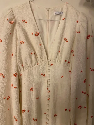 Vit blommig klänning från Adoore - Supersöt vit klänning från Adoore med små röda blommor, v-ringning och dekorativa knappar framtill. Långärmad modell i ett strukturerat tyg som ger en lyxig känsla. Perfekt för sommarens alla tillfällen!. Använd enbart 1gång, det går att lägga bud men går inte under 1350kr