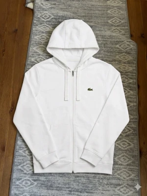 Vit Lacoste Zip-up Hoodie – Classic Fit (Stl S) - Säljer en fräsch och stilren vit luvtröja med dragkedja från Lacoste. Modellen är "Classic Fit" och den lilla klassiska krokodilen är broderad på bröstet. Perfekt basplagg som funkar till allt! 🤍  Storlek: US S (FR 3)  Passform: Classic fit  Färg: Vit Tröjan är i bra begagnat skick. Hör av dig om du har några frågor eller vill samfrakta med något annat i min garderob. Skickas snabbt och spårbart! 📦💨
