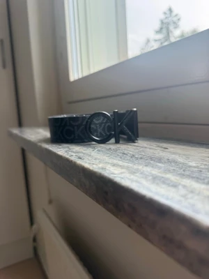 Svart CK bälte med logomönster två sidor - Snyggt svart bälte från Calvin Klein med tydligt CK-logomönster längs hela bandet och en cool metallspänne med CK-detalj. Perfekt accessoar för att lyfta din outfit med en modern touch. Tillverkat i syntetmaterial.
