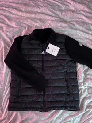 Svart cardiganjacka från Moncler - Snygg svart cardiganjacka från Moncler med quiltad framsida och stickade ärmar. Jackan har dragkedja framtill, hög krage och Moncler-logga på ärmen. Perfekt för lager-på-lager och streetstyle. Säljer eftersom den är för stor gör mig. Aldrig använd. PRIS KAN DISKUTERAS VID SNABB AFFÄR
