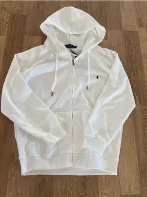 Vit zip hoodie från Polo Ralph Lauren - Snygg vit hoodie med dragkedja från Polo Ralph Lauren. Klassisk design med huva och dragsnören, liten broderad logga på bröstet och två fickor framtill. Perfekt till chill outfits och enkel att matcha med allt.