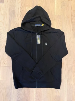 Svart hoodie från Polo Ralph Lauren - Snygg svart hoodie från Polo Ralph Lauren med dragkedja framtill och klassisk vit logga broderad på bröstet. Hoodien har huva med dragsko och ribbade muddar vid ärmslut och nederkant. Perfekt för en avslappnad och stilren look.