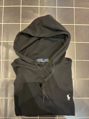 Svart hoodie från Polo Ralph Lauren - Snygg svart hoodie från Polo Ralph Lauren med klassisk huva och dragsko. Ikoniska vita loggan broderad på bröstet. Tillverkad i mjukt bomullsmaterial och har en stor ficka framtill samt ribbade muddar vid ärmar och nederkant. OBS: STORLEKN PÅ TRÖJAN ÄR LITE MINDRE ÄN S FÖR ATT TRÖJAN HAR KRYMT I TVÄTTEN