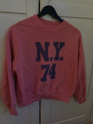 Rosa sweatshirt med N.Y. 74 tryck - Cool rosa sweatshirt med stort mörkblått tryck 'N.Y. 74' på bröstet. Tröjan har rund halsringning, ribbade muddar vid ärmslut och nederkant samt en avslappnad passform. Perfekt för en casual streetstyle-look.