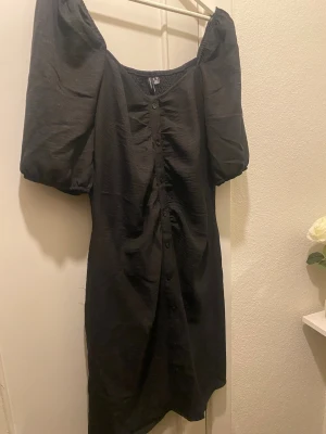 Svart klänning med puffärm Vero Moda - Trendig svart klänning från Vero Moda med puffärmar och knappar framtill. Klänningen har en smockad överdel och en rak passform som ger en snygg siluett. Perfekt för dig som gillar stilrena och moderna plagg.