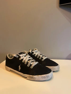 Svarta Polo Ralph Lauren sneakers - Svarta sneakers från Polo Ralph Lauren med vit logga på sidan och vita tjocka skosnören. Skorna har en låg profil och vit sula med slitna detaljer. Klassisk design med rund tå och metallhål för snörning.