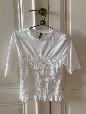 Vit smockad topp från H&M - Supersöt vit topp från H&M med smockad midja och volangkant nedtill. Toppen har rund halsringning och halvlånga ärmar. Perfekt att styla med kjol eller jeans för en fräsch look.