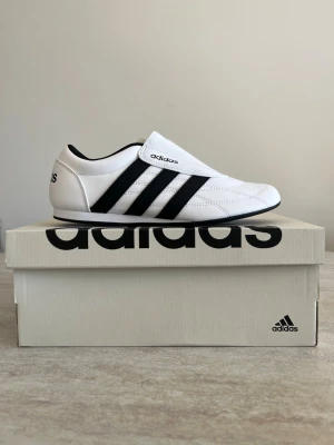 Vita Adidas sneakers med svarta ränder - Snygga vita sneakers från Adidas med klassiska svarta ränder på sidorna och låg profil. Skorna har en slimmad design med kardborrestängning och är tillverkade i syntetmaterial med textildetaljer. Perfekta för en clean och sportig look.