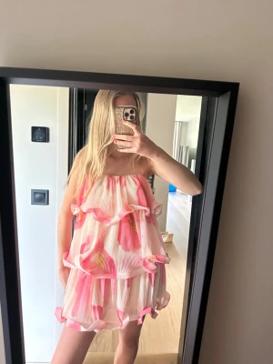 Plisserad volangklänning från NA-KD - Supersöt plisserad miniklänning från NA-KD med volanger och breda axelband. Klänningen har ett rosa och vitt blommigt mönster och är gjord i ett lätt, luftigt material. Perfekt för dig som gillar statement-plagg och vill sticka ut med färg och form.