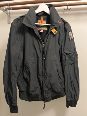 Svart bomberjacka från Parajumpers - Snygg svart bomberjacka från Parajumpers med patch på ärmen och coola detaljer som orange dragkedja och märkestag. Jackan har flera fickor, ribbade muddar och är tillverkad i lätt nylonmaterial. Perfekt för dig som vill ha en streetig och trendig look.