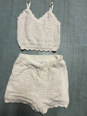 Vitt stickat set med topp och shorts - Sött vitt stickat set med en croppad topp med smala axelband och vågig kant, samt matchande shorts med bred resår i midjan och vågig kant nedtill. Setet har ett diskret hålmönster och känns både mjukt och luftigt. 