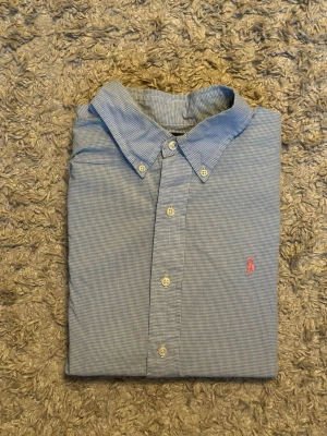 Blå rutig skjorta från Ralph Lauren - Klassisk blå rutig skjorta från Ralph Lauren med knappkrage och lång ärm. Skjortan har små vita knappar framtill och en broderad rosa logga på bröstet. Tillverkad i mjuk bomull som känns skön mot huden. Perfekt för en clean och stilren look.