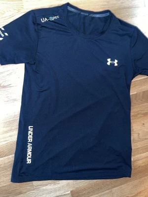 Mörkblå träningströja Under Armour - Snygg mörkblå träningströja från Under Armour med rund hals och korta ärmar. Tröjan har vita logotyper på bröstet och sidan samt texten UA-SPEC på axeln. Tillverkad i ett lätt och snabbtorkande funktionsmaterial, perfekt för löpning eller gym.