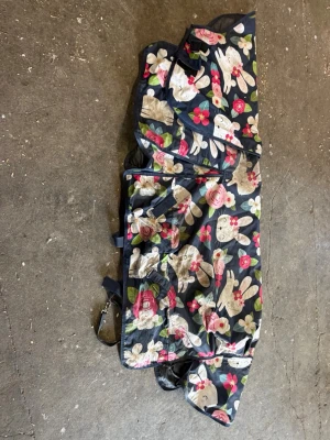 Blommig väska med kaninmotiv - Säljer ett hästtäcke med mörkblå botten och blommigt mönster i rosa, vitt och grönt. Täcket har tryck av kaniner och flera spännen för att fästa runt hästen. Materialet känns som slitstark syntet och täcket är ett Vintertäcke med 250g och storlek 125cm 