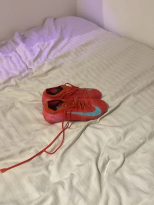 Nike röda fotbollsskor med blå swoosh - Säljer ett par snygga fotbollsskor från Nike i röd färg med en stor blå swoosh på sidan. Skorna har klassisk snörning och dobbar för bra grepp på planen. Tillverkade i syntetmaterial för lätt känsla och bra bollkontroll. Perfekta för dig som vill sticka ut på fotbollsplanen.