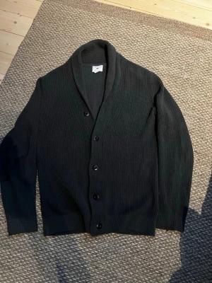 Svart ribbad kofta från H&M i st S - Säljer en svart ribbad kofta från H&M med sjalkrage och knäppning framtill. Koftan har långa ärmar och en klassisk, stilren look som passar perfekt till lager-på-lager. Materialet är mjukt och bekvämt, perfekt för kyligare dagar. Storleken är igentligen xs men sitter som s skulle jag säga
