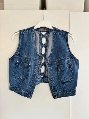 Blå jeansväst med snörning bak - Säljer en blå jeansväst i klassisk denim med snygg snörning i ryggen och två stora fickor framtill. Västen har metallknappar och coola sömdetaljer som ger en retro känsla. Perfekt för att lyfta vilken outfit som helst! Ingen storlek i men skulle säga en s