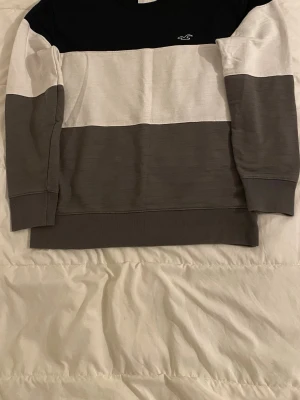 Randig sweatshirt från Hollister.  - Snygg sweatshirt från Hollister med breda blockränder i svart, vitt och grått. Tröjan har rund halsringning, långa ärmar och en liten broderad logga på bröstet. Perfekt för en avslappnad och stilren look.