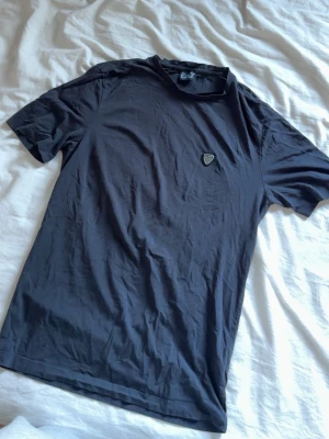 Svart t-shirt från EA7 Emporio Armani - Snygg svart t-shirt från EA7 Emporio Armani med diskret logga på bröstet. Klassisk passform och rund halsringning. Tillverkad i mjuk bomull som känns skön mot huden. Perfekt till jeans eller shorts för en clean look.