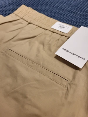 Beige shorts från These Glory Days 58 XXl - Stilrena beige shorts från These Glory Days med elastisk midja och diskret bakficka. Klassisk rak passform och tillverkade i mjuk bomull, perfekta för varma dagar. Enkla att matcha med t-shirt eller skjorta för en clean look.