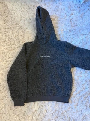 Mörkgrå teddyhoodie Originals Studio - Mysig mörkgrå teddyhoodie från Originals Studio/ Jack and Jones med broderad logga på bröstet. Hoodien har en stor huva, ribbade muddar vid ärmslut och nederkant samt är gjord i fluffigt teddy-material som håller dig varm. Perfekt för chill dagar eller när du vill vara extra bekväm. Nypris 579kr. Herr storlek.