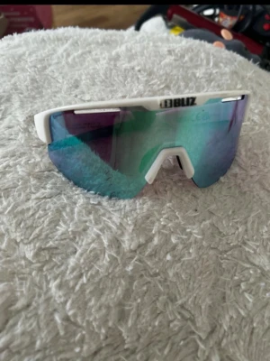 Vita sportiga solglasögon från Bliz - Säljer ett par vita sportiga solglasögon från Bliz med spegelglas i blå och lila toner. Modellen har en modern, kantig form och breda skalmar med tydlig logga. Perfekta för dig som vill ha en cool och funktionell look.