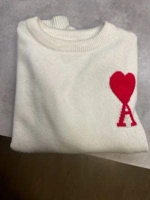 Vit stickad tröja från Ami Paris - Säljer en vit stickad tröja från Ami Paris med röd hjärtlogga och bokstaven A på bröstet. Tröjan har rund halsringning och ribbade muddar. Perfekt för dig som gillar stilrena plagg med en twist.