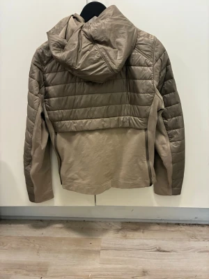 Beige pufferjacka med huva - Snygg beige pufferjacka med quiltade partier och dragkedja framtill. Jackan har två fickor med dragkedja och en fast huva. Har en modern, avslappnad look. Materialet är troligtvis syntet med vaddering.