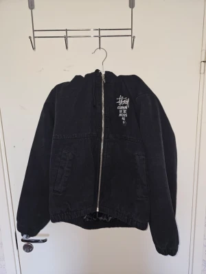 Svart Stüssy jacka med dragkedja - Svart jacka från Stüssy med vit broderad logga och text på bröstet. Jackan har dragkedja framtill, två sidofickor och resår vid ärmslut och midja. Materialet är kraftigt tyg, troligen bomull, och modellen är avslappnad och streetinspirerad.