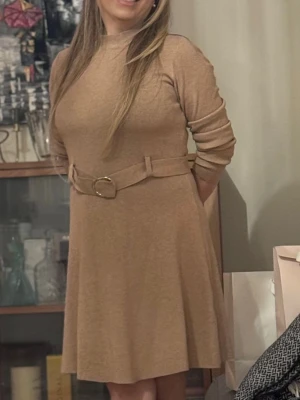 Beige klänning med bälte - Snygg beige klänning med lång ärm och matchande bälte i midjan. Klänningen har en mjuk, lätt utställd passform och rund halsringning. Perfekt för dig som gillar stilrena och enkla plagg med en twist.