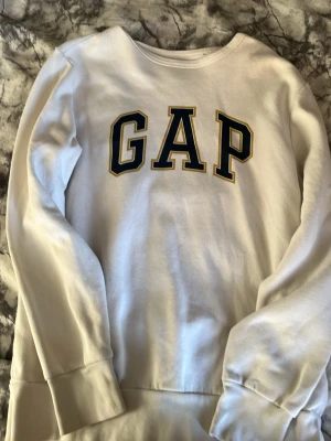 Vit GAP hoodie med tryck - Vit hoodie från GAP med klassiskt stort GAP-tryck i mörkblått och gult på bröstet. Mjuk insida och ribbade muddar vid ärmslut och nederkant. Perfekt för en avslappnad och stilren look.