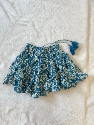 Blåvit kortkjol med volanger och tofsar - Kort kjol i blått och vitt med volanger och elastisk midja. Kjolen har ett fint mönster och dekorativa knytband med tofsar. Tillverkad i ett lätt och luftigt bomullstyg. Kjolen ör från Gina Young men storleken motsvarar xxs. Fråga för mer bilder!