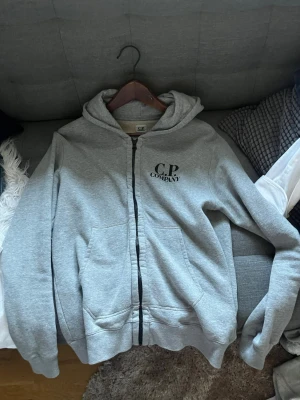 Grå hoodie från C.P. Company - Snygg grå hoodie från C.P. Company med dragkedja framtill och svart logga på bröstet. Hoodien har två fickor och en klassisk huva. Tillverkad i mjukt sweatshirtmaterial som är perfekt för chill dagar.