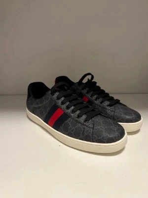 Gucci Ace sneakers med GG-mönster - Snygga Gucci Ace sneakers i svart med GG-monogram, röda och blå ränder på sidan samt kontrasterande häl i rött och blått med metallic-finish. Skorna har vita sulor och svarta skosnören. Perfekta för dig som gillar exklusiv streetstyle. Det är storlek 45 men passar även 44 då dem är små i storlek