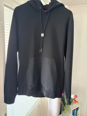 Svart hoodie från Hugo Boss - Snygg svart hoodie från Hugo Boss med klassisk huva och dragsnören med metalländar där det står HUGO. Stor känguruficka framtill i ett glansigare material som ger en cool kontrast. Perfekt för dig som gillar stilrena och moderna plagg.