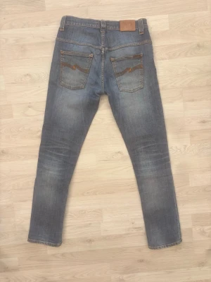 Nudie Jeans - Säljer ett par klassiska blå slim fit jeans från Nudie Jeans. Jeansen har fem fickor, slitningar och snygga detaljer på bakfickorna. Tillverkade i mjuk denim med en tvättad look och kontrastsömmar. Perfekta för en avslappnad och trendig stil.
