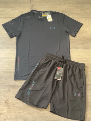 Under Armour svart träningsset - Säljer ett svart träningsset från Under Armour med t-shirt och shorts. Båda plaggen har logotypen tryckt i färg och är tillverkade i lätt och snabbtorkande material. Perfekt för gymmet eller löpning. Snygg design med diskreta detaljer och elastisk midja på shortsen.