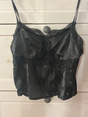 Svart satin linne med spetsdetaljer - Snygg svart topp i satin med tunna axelband och dekorativa spetsdetaljer. Toppen har en dragkedja på sidan och ett coolt cut-out parti framtill som ger en edgy look. Perfekt att styla med jeans eller kjol för en trendig outfit.